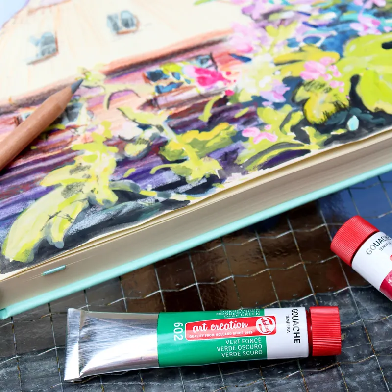 Tubka zielonej farby gwaszowej Talens Art Creation obok szkicownika z malowanym pejzażem.
